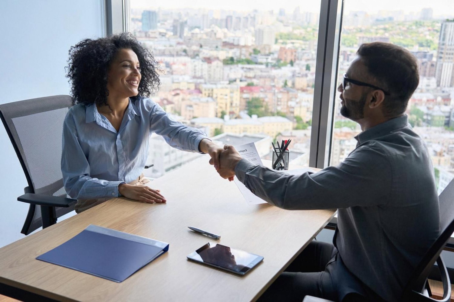 Dubai employer and employee shaking hands across a desk during an HR meeting with a city skyline behind them
